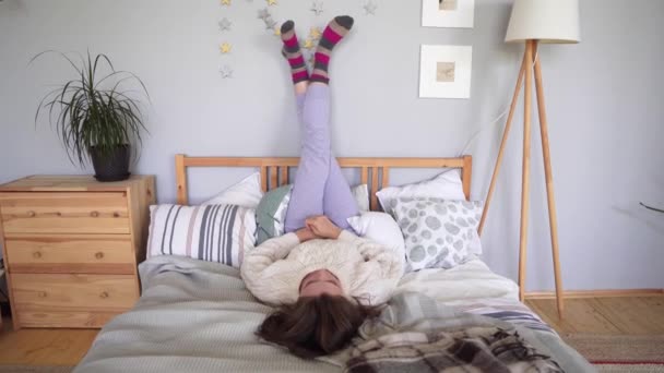 A cheerful young woman lies on a bed with her legs on the wall, fooling around and resting in home clothes and colorful socks — Stock Video