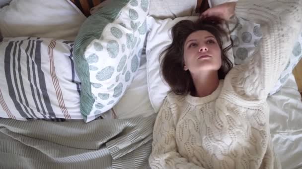 白いセーターを着た若い魅力的な女性はベッドに横たまり、夢を見てベッドに入る — ストック動画