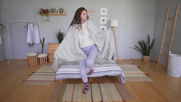 Mooie jonge vrouw in pyjama en een trui is dansen en plezier naast het bed, verbergen in een deken in de slaapkamer. Hygge of Lagom concept — Stockvideo