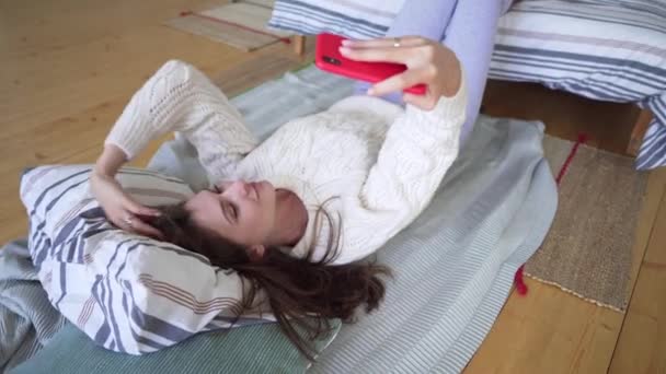 Menina atraente encontra-se em um cobertor perto da cama e toma uma selfie no telefone, brincando e bocejando . — Vídeo de Stock