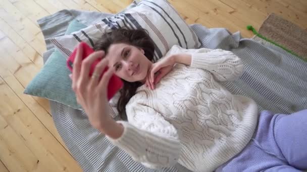 Bella donna caucasica in un maglione bianco a maglia prende un selfie mentre si trova su un plaid accogliente e cuscino nella sua camera da letto — Video Stock