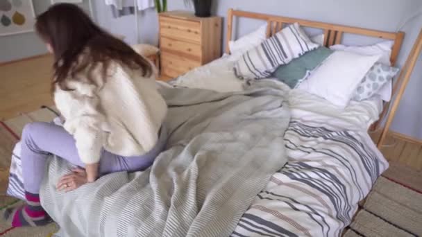 Μια γυναίκα με ρούχα στο σπίτι σηκώνεται από το κρεβάτι και φτιάχνει το κρεβάτι με ένα γκρίζο καρό. Loft διαμέρισμα σκανδιναβικού στυλ — Αρχείο Βίντεο