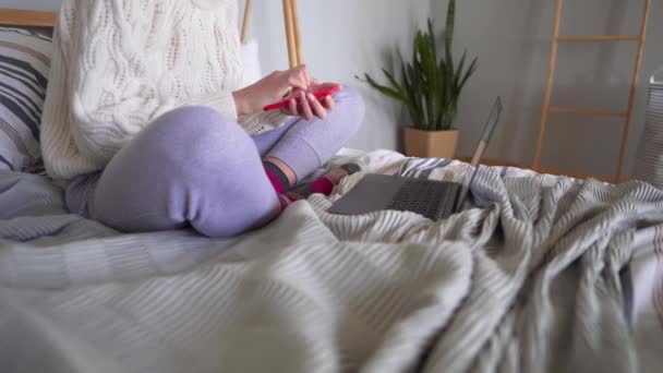 Крупный план - женщина использует телефон и ноутбук для работы из дома. Фрилансер, сидящий в пижаме в постели — стоковое видео