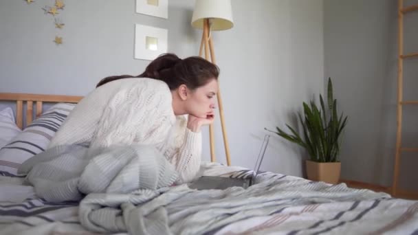 Mujer cansada freelancer trabajando en un portátil en una casa fría, sentada en un suéter y cálida pijama de casa en la cama. Estrés y desventajas del trabajo por cuenta propia — Vídeo de stock