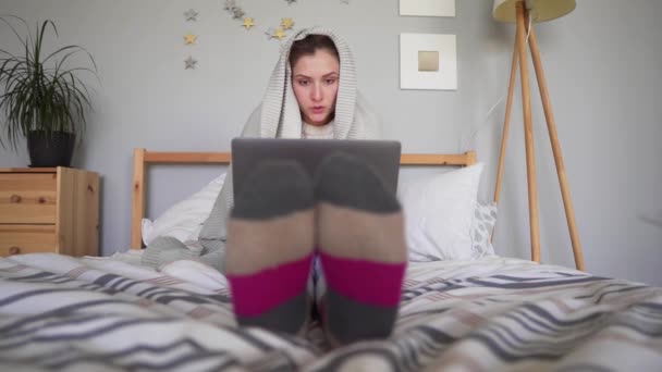 Una joven con calcetines de color rosa grisáceo trabaja en una computadora portátil, envuelta en una manta a cuadros, sentada en la cama en casa . — Vídeos de Stock