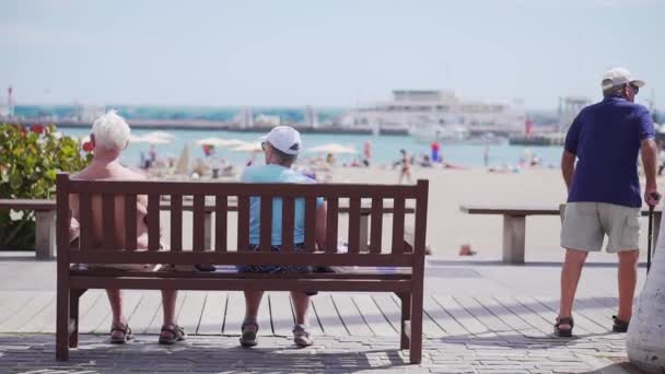 Los Cristianos, Spanien-april, 17, 2019-äldre pensionerade på semester. Pensionärer sitter på en bänk på havet, framför en sandstrand, människor passerar genom. — Stockvideo