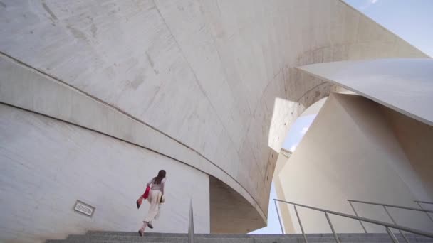 Una chica hipster con un sombrero en la mano se levanta por las escaleras y se dirige a un hermoso edificio moderno: el Auditorio de Tenerife. Turista mujer caucásica en la atracción principal de la ciudad, arquitectura de arte — Vídeos de Stock