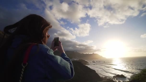 Joven mujer hipster fotografía por teléfono el impresionante paisaje de la costa al atardecer. Viento soplando pelo. Paisaje marino canario, paisaje océano — Vídeos de Stock