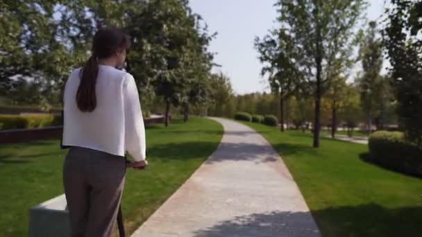 추적 샷 - 흰색 셔츠를 입은 어린 소녀가 따뜻한 여름날에 녹색 공원에서 타고 있습니다. 교통의 트렌디 한 에코 모드 — 비디오