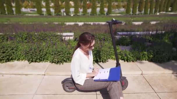 Mujer trabajadora desarrolla tecnología en un cuaderno, sentada en un scooter eléctrico en un hermoso parque en un día soleado de verano . — Vídeos de Stock