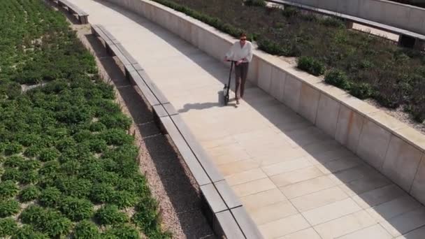 Atractiva mujer caucásica está caminando con un scooter en una hermosa ciudad moderna en un día soleado, vista aérea. Moderno transporte ecológico, popular entre los jóvenes — Vídeo de stock