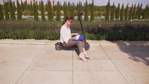 Joven mujer bonita sentada en un scooter eléctrico en un soleado día de verano en un parque. Estudiante crear con bloc de notas al aire libre — Vídeo de stock