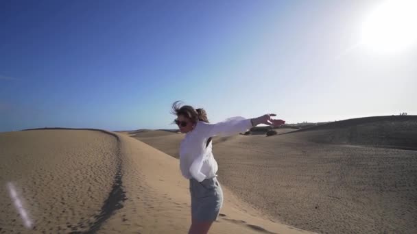 Jovem atraente com uma camisa branca e óculos no deserto em uma duna gira em torno de si mesma. Diversão em férias ao ar livre — Vídeo de Stock