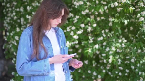 一个漂亮的女孩通过扫描手机上的qr代码把卡片和手机绑在一起。一个女人，在街上拿着电话，背靠着花 — 图库视频影像