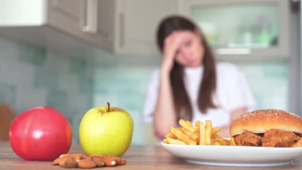 Η επιλογή μεταξύ ανθυγιεινών και υγιεινών τροφίμων. Μπέργκερ και τηγανητές πατάτες μπροστά από ένα μήλο, ντομάτα και καρύδι. Λεπτή κοπέλα στην κουζίνα προσπαθεί να κάνει μια δύσκολη επιλογή των τροφίμων — Αρχείο Βίντεο