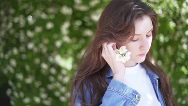 Vrouw bloemblaadje. Aantrekkelijk meisje zet achter zijn oor een tak van jasmijn met witte geurige bloemen en glimlachen. Vrouwenportret in de natuur tegen de achtergrond van struiken — Stockvideo