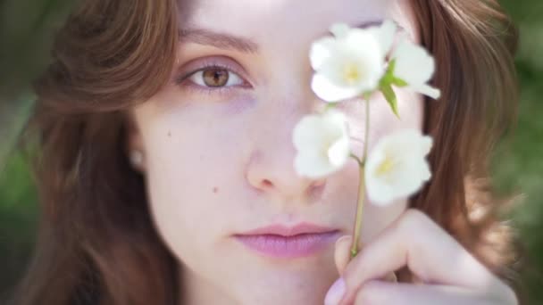 Portrait d'une jeune femme en gros plan, couvrant son œil d'une branche de jasmin. Belle fille souriante avec des fleurs à la main — Video