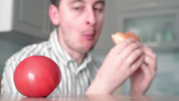 Der Mann wählte einen ungesunden Lebensstil. Guy isst Burger im Hintergrund frische Tomaten — Stockvideo