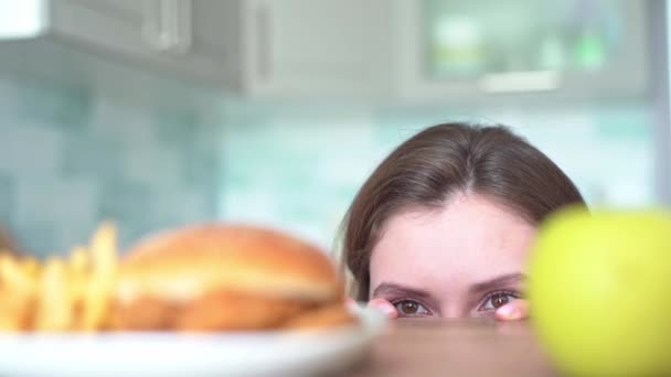 Womens eyes look around choosing between harmful fasfood and a healthy fruit. Dilemma of choice from a young woman between a burger with fried fries and an apple — Stock Video