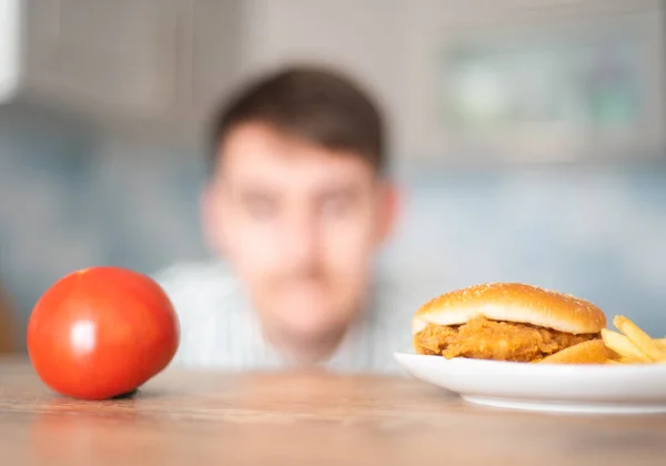 Голодний чоловік вибирає між сирником і помідором. Швидка їжа або овочева дієта. Спокуса нездорової їжі — стокове фото