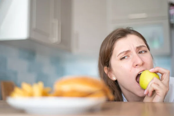 Wilskracht op dieet. De juiste keuze - een vrouw eet een gezonde appel tegen de achtergrond van schadelijk fastfood — Stockfoto