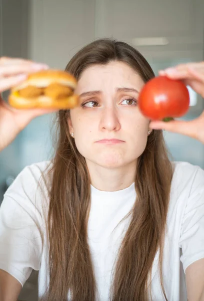 Verleiding van junkfood. Het meisje denkt na over wat ze moet eten. Een mooie vrouw houdt een tomaat en cheeseburger vast en kijkt ernaar. — Stockfoto