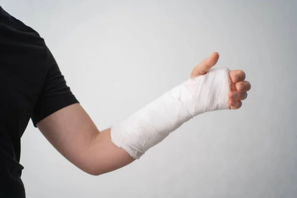 A young man shows the injury of a broken arm in a plaster bandage on a white background. The mans fracture hand barely flexes his fingers