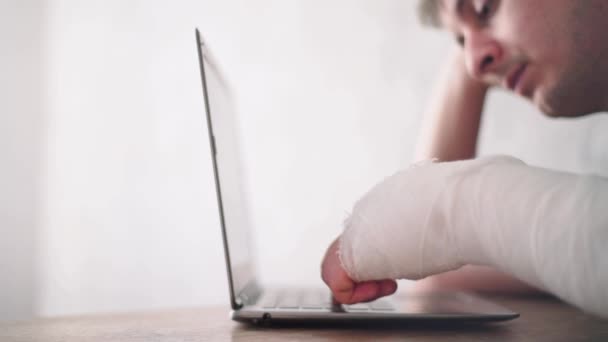 Um homem com uma lesão no braço olha para um portátil. Diagnóstico online. Seguro de saúde — Vídeo de Stock