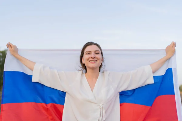 Mulher russa bonita com bandeira russa em suas mãos, retrato, vista frontal. A mulher levanta a bandeira pelas costas, sorri e olha para a câmera — Fotografia de Stock
