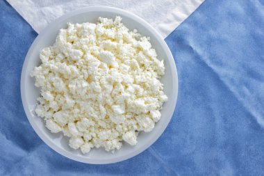 Bir beyaz mavi kumaş arka plan, beyaz bir plaka üzerinde yumuşak peynir, taze ev yapımı peynir beyaz mavi peçeteye süzme peynir. Süt ürünleri vintage tarzı, sağlıklı bir yaşam tarzı. Kopya alanı