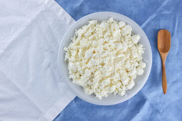 Cottage cheese on a white blue fabric background, soft cheese on a white plate, fresh homemade cheese on a white blue napkin. Dairy product in vintage style, healthy lifestyle. Copy space