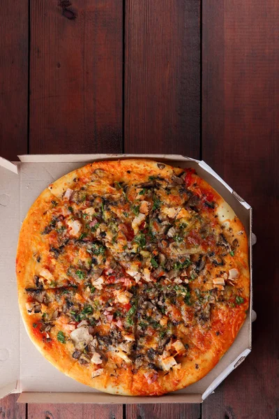 Pizza in a box on a wooden background. Italian pizza with chicken, mushrooms, greens, cheese and sauce. Festive meal in a cardboard box