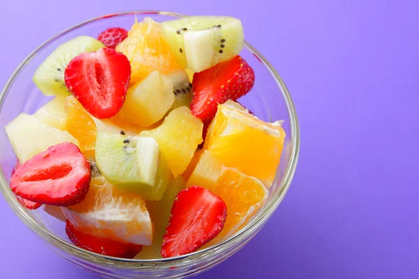 Fruit salad from pineapple, kiwi, orange, strawberry. Multicolored sliced fruit in a transparent dish. Vegetarian food on a violet background. Copy space