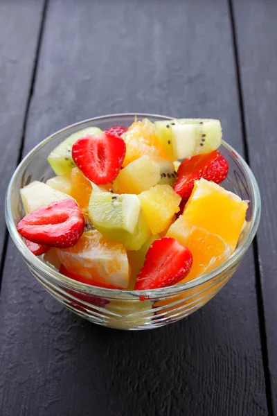 Fruit salad from pineapple, kiwi, orange, strawberry. Multicolored sliced fruit in a transparent dish. Vegetarian food on a black wooden background
