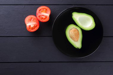 Avokado ve siyah bir tabakta domates domates bir ahşap arka plan siyah, kırmızı ve yeşil sebze üstten görünüm, minimalist tarzda egzotik meyve ve avokado yarısını