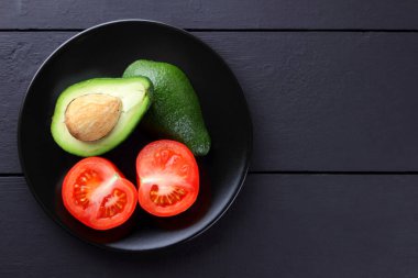 Avokado ve siyah bir tabakta domates domates bir ahşap arka plan siyah, kırmızı ve yeşil sebze üstten görünüm, minimalist tarzda egzotik meyve ve avokado yarısını