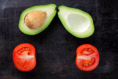 Avokado ve karanlık bir metal yüzeyi domates Yarım avokado ve yaşlı bir metal arka plan, kırmızı ve yeşil sebze yukarıdan, minimalist tarzda egzotik meyve domates