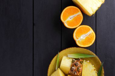 Ananas ve ahşap panoları, ananas meyve ve portakal karanlık bir arka plan, tropikal meyve salatası kahvaltıda vejetaryen yemek, üzerinde sarı bir plaka üzerinde yarım portakal parçaları alanı Kopyala