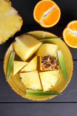 Ananas ve ahşap panoları, ananas meyve ve portakal karanlık bir arka plan, tropikal meyve salatası kahvaltıda vejetaryen yemek, üzerinde sarı bir plaka üzerinde yarım portakal parçaları alanı Kopyala