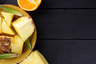 Ananas ve ahşap panoları, ananas meyve ve portakal karanlık bir arka plan, tropikal meyve salatası kahvaltıda vejetaryen yemek, üzerinde sarı bir plaka üzerinde yarım portakal parçaları alanı Kopyala