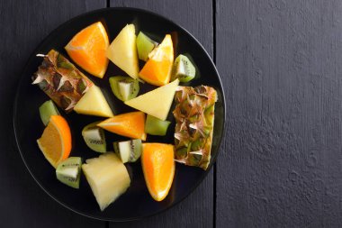 Ahşap panoları, ananas, portakal ve kivi karanlık bir arka plan üzerinde Kahvaltı, vejetaryen yemek, kopya alanı için tropikal meyve salatası üzerinde siyah bir plaka üzerinde meyve salatası