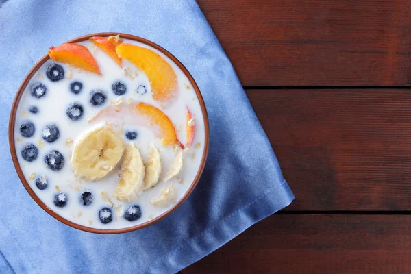 Oatmeal porridge with milk and fruits on a blue napkin, blueberry on wooden boards, vegetarian food on textiles, useful porridge with pieces of banana and peach