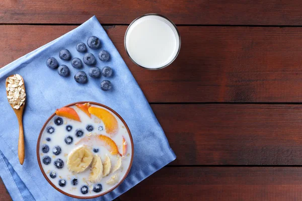 Oatmeal porridge with milk and fruits on a blue napkin, blueberry on wooden boards, vegetarian food on textiles, useful porridge with pieces of banana and peach, glass with milk