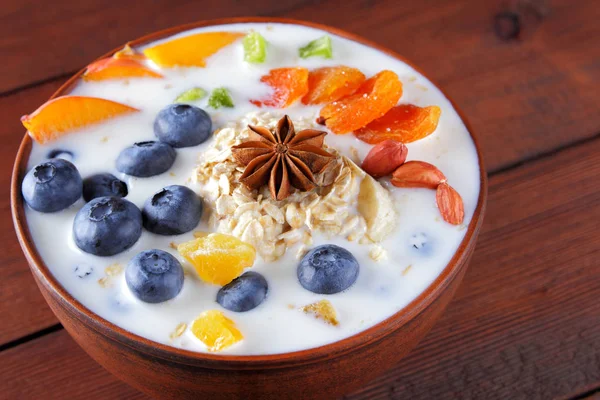 Oatmeal porridge with milk, fruits and peanuts on wooden background, healthy porridge with banana and peach slices, breakfast with fiber, calcium and vitamins, anise, candied fruits and dried apricots in milk