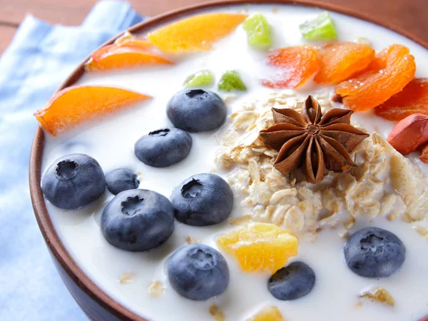 Oatmeal porridge with milk, fruits and peanuts on wooden background, healthy porridge with blueberries, banana and peach slices, breakfast with fiber, food on blue napkin