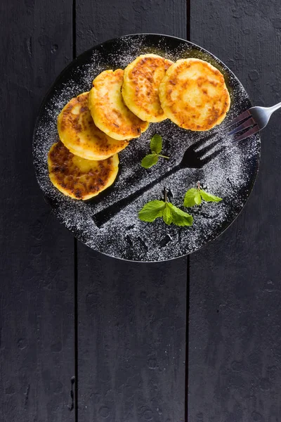 Cottage cheese pancakes on a dark background. Traditional Ukrainian dish - syrniki with fresh mint. Pancakes with cottage cheese on a black plate sprinkled with powdered sugar. Copy space