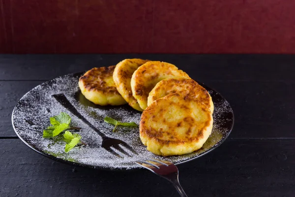Cottage cheese pancakes on a dark background. Traditional Ukrainian dish - syrniki with fresh mint. Pancakes with cottage cheese on a black plate sprinkled with powdered sugar. Copy space