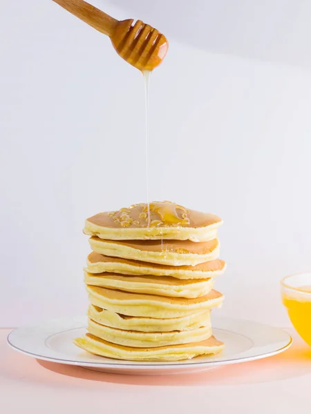 Pancakes on a white pink background. Hot pancakes with honey on a white plate with copy space. Delicious dish for breakfast in the style of pop art. Honey flows down from a wooden stick on a dish