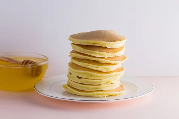 Pancakes on a white pink background. Hot pancakes with honey on a white plate with copy space. Delicious dish for breakfast in the style of pop art. Honey flows down from a wooden stick on a dish