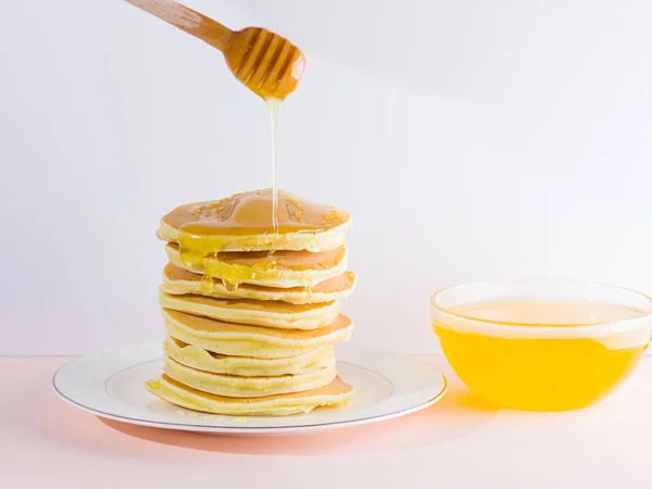 Pancakes on a white pink background. Hot pancakes with honey on a white plate with copy space. Delicious dish for breakfast in the style of pop art. Honey flows down from a wooden stick on a dish
